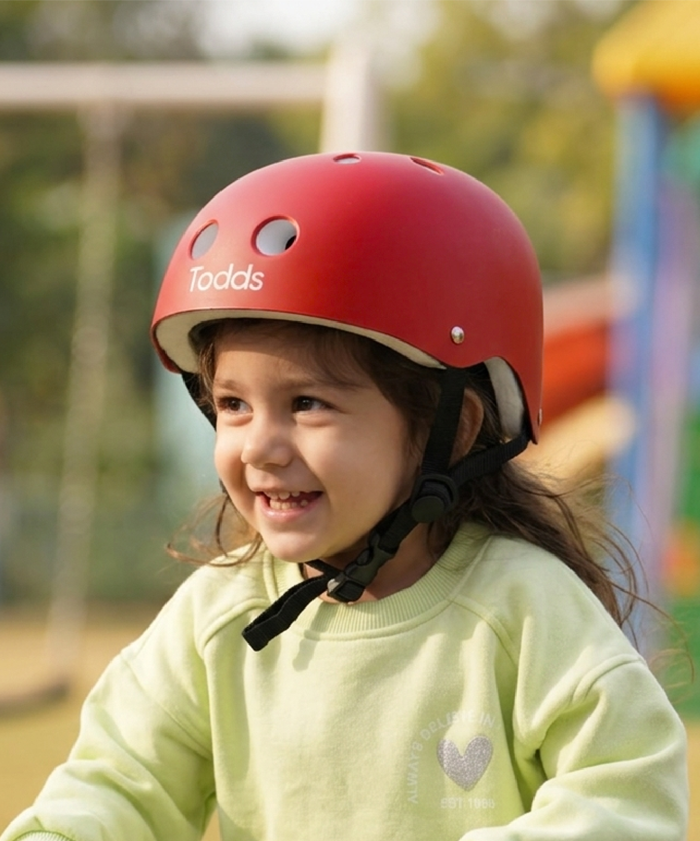 A young child riding a green scooter in a playground, wearing a red toddler helmet with the brand name 'Toddsl' and a pale yellow-green sweatshirt and matching pants.